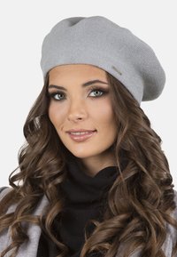 Boina gris de punto con forma redonda y flexible. Incluye un pequeño acento metálico. Se lleva sobre el cabello rizado y suelto con un cuello alto negro.