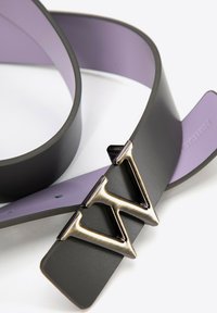 Cinturón reversible en negro y lavanda que presenta una hebilla de metal con un logo estilizado. El cinturón tiene una textura suave y un diseño minimalista.