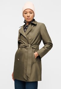 Γυναίκα που φοράει μπεζ πλεκτό καπέλο, χακί παλτό τύπου trench με ζώνη και μπλε τζιν, στέκεται μπροστά σε απλό λευκό φόντο.