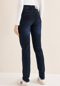 Jeans de mezclilla azul oscuro con cintura alta, corte de pierna recta y dos bolsillos traseros. Combinados con zapatillas blancas con detalles en rojo.
