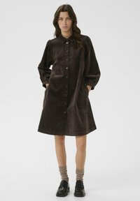 Brown corduroy button-up dress with a collared neck, long puffed sleeves, and side pockets, falling above the knees.