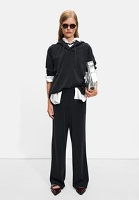 Black oversized hoodie with drawstrings, layered over a white shirt, paired with wide-leg black trousers and pointed burgundy loafers. Silver bag.