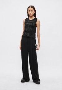 Črna brezrokavnik z navpično pleteno teksturo, kombiniran s širokimi črnimi hlačami in drzno platformno obutvijo, z metalnim usnjenim torbico v roki.