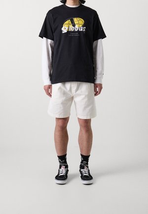 Camiseta gráfica negra con diseño de globo amarillo, combinada con pantalones cortos blancos, zapatillas negras y calcetines negros con estampado. Ajuste casual y relajado.