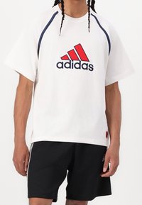 Camiseta blanca con acentos en azul marino y rojo, que presenta un gran logo de Adidas en rojo y negro, cuello redondo y mangas cortas, combinada con pantalones cortos negros.