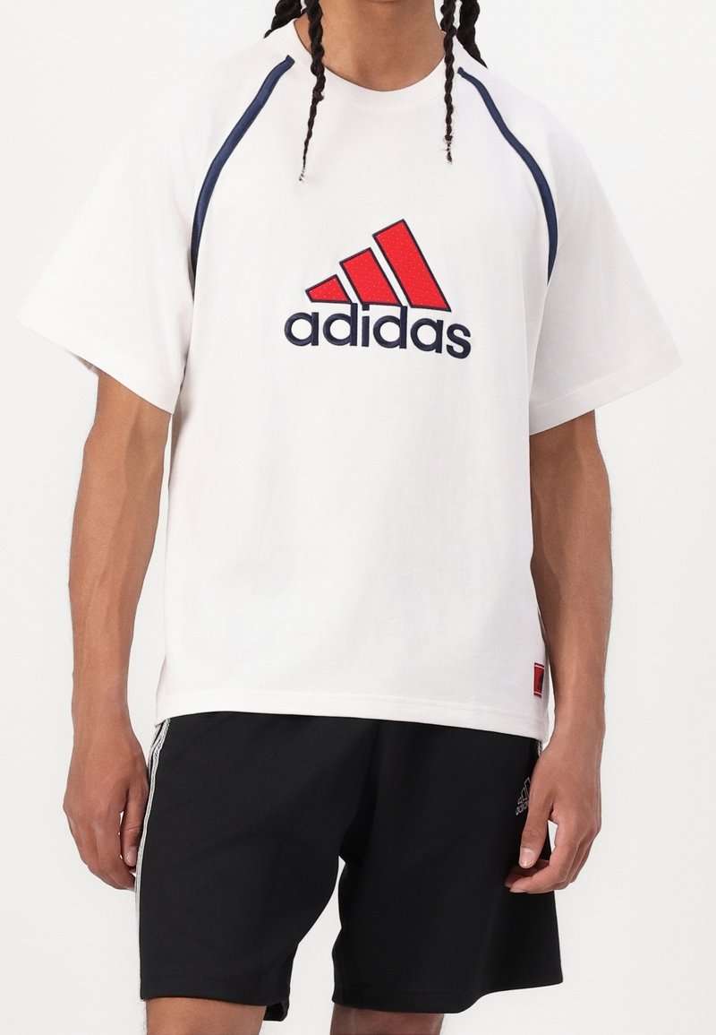 Camiseta blanca con acentos en azul marino y rojo, que presenta un gran logo de Adidas en rojo y negro, cuello redondo y mangas cortas, combinada con pantalones cortos negros.