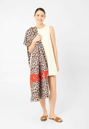 Frau in ärmellosem cremefarbenem Kleid, die einen Stoff mit Leopardenmuster und großem roten Text hält, trägt braune Sandalen und steht vor weißem Hintergrund.