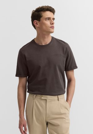 Mann trägt ein dunkelbraunes T-Shirt mit kurzen Ärmeln und beigen Hosen, schaut nach rechts vor einem schlichten hellen Hintergrund.