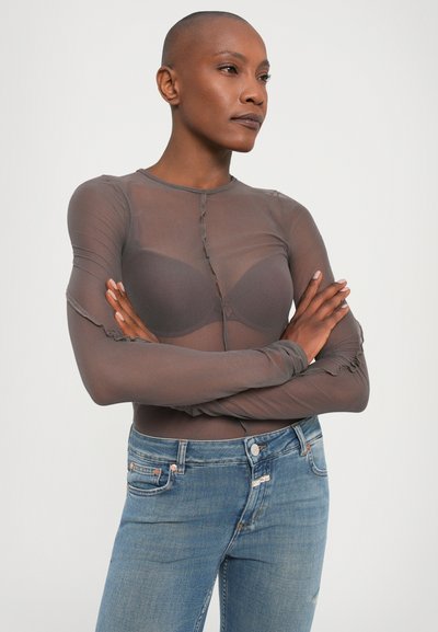Transparante bruine top met lange mouwen en een ronde halslijn, knoopsluiting en geplooide mouwdetails, gecombineerd met lichtblauwe strakke jeans.