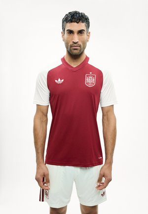 Uomo in piedi rivolto in avanti che indossa la maglia della nazionale di calcio spagnola con torso rosso, maniche bianche e pantaloncini bianchi su sfondo bianco.