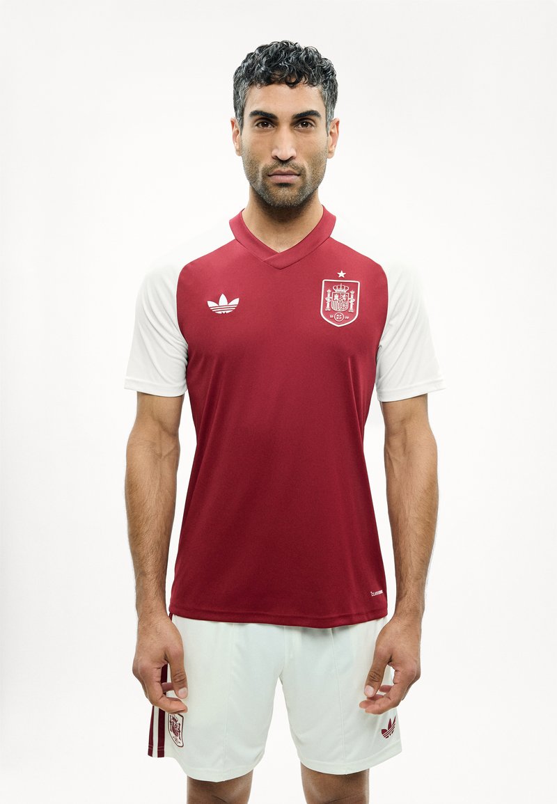 Mand står vendt mod fronten iført en spansk fodboldtrøje med rød torso, hvide ærmer og hvide shorts mod hvid baggrund.