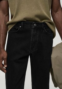 Pantalones vaqueros de mezclilla negra con un corte recto, con bragueta de botones, bolsillos delanteros y detalles de costura sutiles. Combinados con una camisa verde oliva.