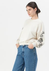 Felpa color crema con motivi floreali neri sulle maniche, polsini a coste e orlo. Abbinata a jeans blu chiaro per un look rilassato.