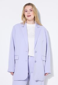 Blazer oversized en lila con solapas de muesca, dos botones, bolsillos laterales y una textura suave; combinado con una blusa blanca sencilla.