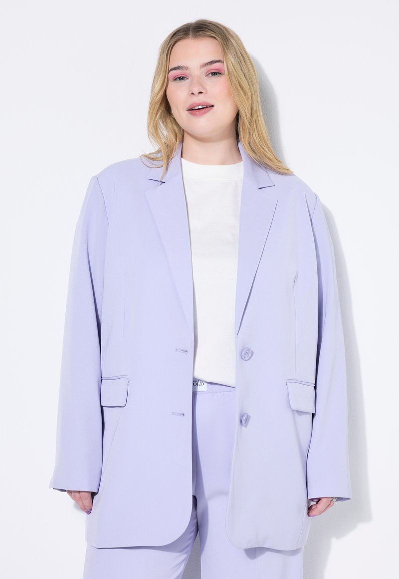 Blazer oversized en lila con solapas de muesca, dos botones, bolsillos laterales y una textura suave; combinado con una blusa blanca sencilla.