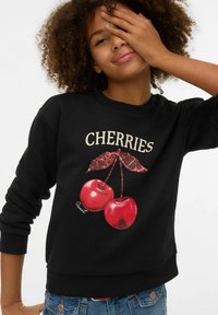 Melns džemperis ar sarkanu ķiršu grafiku un zaļām lapām, uz kura ir gaišiem burtiem rakstīts vārds "CHERRIES". Mīksts audums, ar ribotām aprocēm.