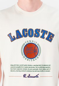 Krēmpadānas kokvilnas T-krekls ar izteiksmīgu sarkanu un zilu uzrakstu "LACOSTE" un "TENNIS HERITAGE", ar zaļu un oranžu krokodila logo.