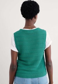 Green knitted vest with textured pattern, featuring wide armholes and a ribbed hem, paired with a white t-shirt and blue jeans.