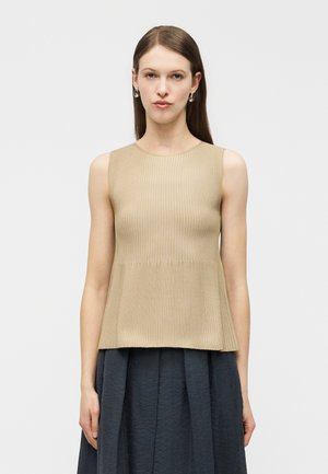 Femme portant un haut beige côtelé sans manches et une jupe plissée foncée, posant devant un fond clair uni.