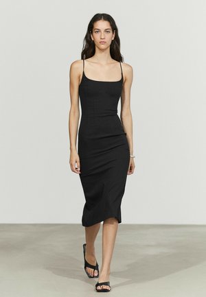 Frau läuft, trägt ein figurbetontes schwarzes Midikleid mit Spaghettiträgern und schwarze offene High Heels vor einem einfarbigen hellen Hintergrund.