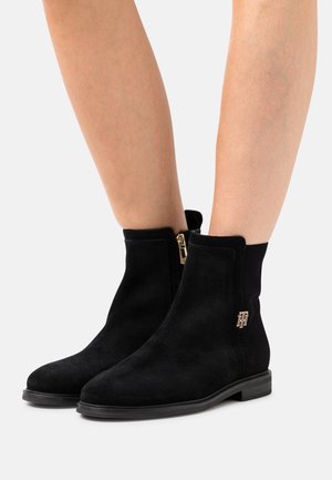 Černé semišové kotníkové boty se zipem na boku, plochou gumovou podrážkou a zlatým logem na boku. Texturovaný povrch a elegantní design.