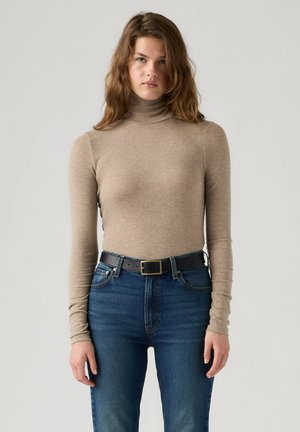 Ung kvinde med bølget brunt hår iført en beige højhalset trøje og blå jeans med sort bælte står foran en ensfarvet baggrund.