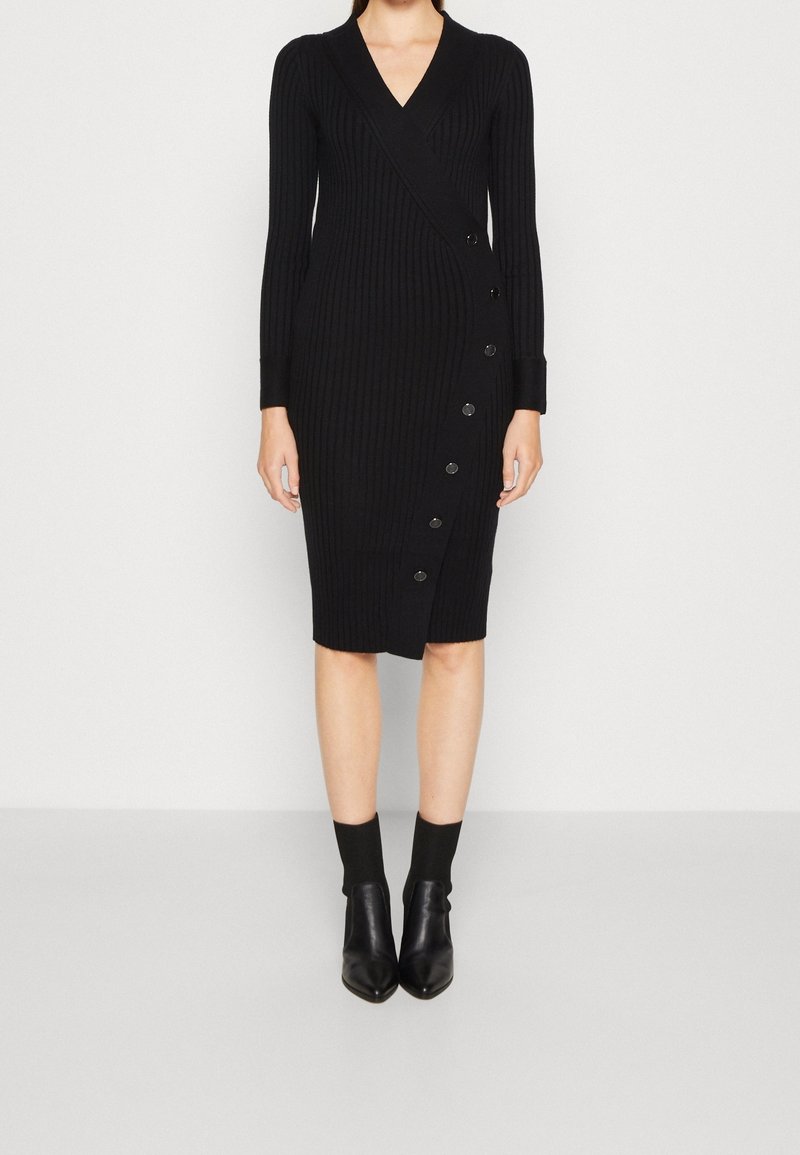 Robe portefeuille noire en maille avec manches longues, col en V, texture côtelée et huit boutons décoratifs sur le côté. Longueur jusqu'aux chevilles.