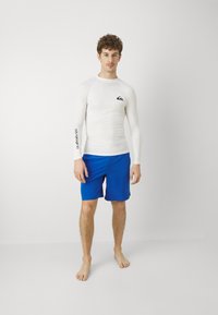 Długi rękaw biały rash guard z czarnym logo na klatce piersiowej, w połączeniu z jasnoniebieskimi szortami kąpielowymi, bose stopy, stojący na neutralnym tle.