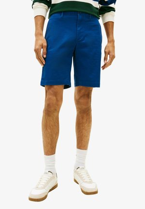 Homme portant un short bleu vif jusqu'aux genoux, des chaussettes blanches et des baskets beiges, debout les bras détendus le long du corps.