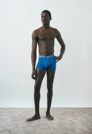 Λεπτός, χωρίς πουκάμισο άνδρας στέκεται ξυπόλητος πάνω σε χαλί, φοράει μπλε εσώρουχα μπόξερ της Nike, με το ένα χέρι στη μέση και ουδέτερη έκφραση.