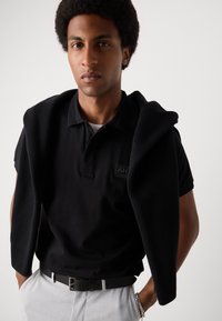 Zwarte polo met korte mouwen en een ribgebreide kraag, met een logo op de patch. Een zwarte gebreide trui ligt over de schouders. Lichtgekleurde broek.