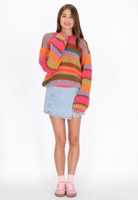 Dzianinowy, oversize'owy sweter w wielobarwne paski w kolorach pomarańczowym, różowym, zielonym i fioletowym, zestawiony z jasnoniebieską mini spódnicą dżinsową i różowymi butami.