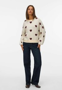 Femme portant un pull blanc avec des motifs de cœur bourgogne, un jean bleu foncé et des chaussures brunes, se tenant devant un fond blanc uni.