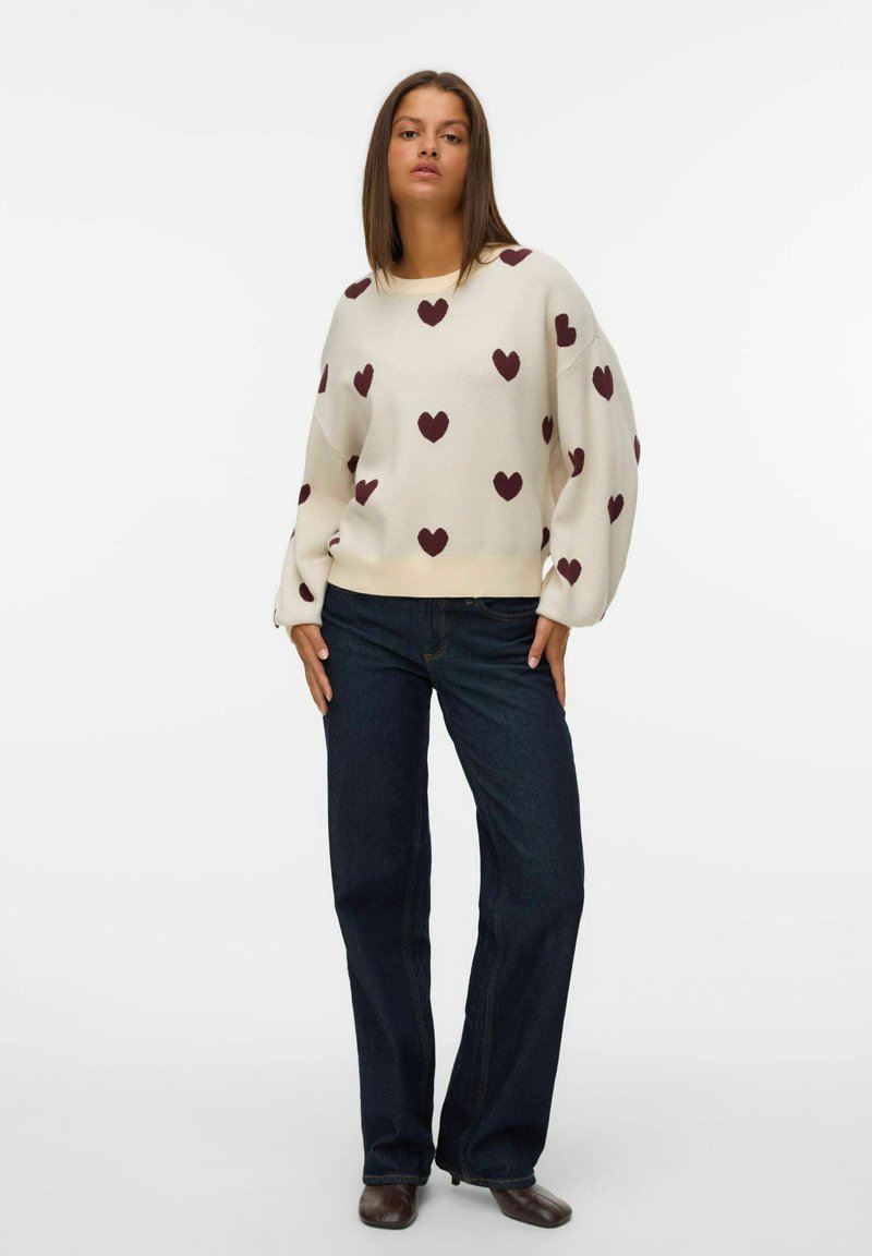 Femme portant un pull blanc avec des motifs de cœur bourgogne, un jean bleu foncé et des chaussures brunes, se tenant devant un fond blanc uni.