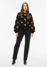 Czarny dziany sweter z beżowymi wzorami serc, okrągłym dekoltem i rękawami typu balloon, zestawiony z czarnymi spodniami i botkami za kostkę.