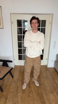 Νέος άνδρας με καστανά μαλλιά που φοράει λευκό πλεκτό πουλόβερ, μπεζ παντελόνι cargo και ανοιχτά αθλητικά παπούτσια, στέκεται με τα χέρια σταυρωμένα στην είσοδο του σαλονιού.