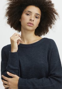 Ciemnoniebieski sweter dziany z szerokim dekoltem, ściągaczami na rękawach i miękką teksturą. Modelka pozuje z założonymi na siebie rękami, eksponując detale ubrania.