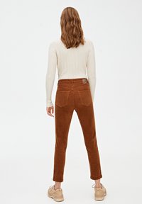 Femme aux cheveux bruns ondulés portant un pull en maille torsadée blanc, un pantalon en velours côtelé marron et des baskets beiges, debout devant un fond uni.