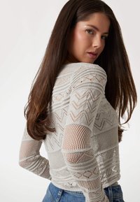 Pulover din tricotaje gri deschis, cu modele lucrate la broderie deschisă, prezentând texturi în zigzag și plasă pe mâneci și corp, cu decolteu rotund.
