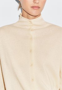 Beige, stickad kofta med hög krage och sex matchande knappar. Mjuk textur med ett lätt ribbat mönster.