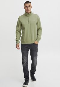 Sudadera verde de media cremallera con una textura suave, que presenta un pequeño logo, combinada con jeans oscuros y zapatillas negras.