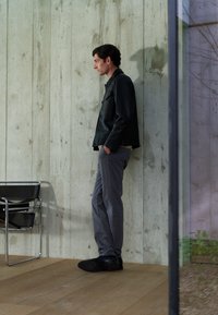 Zwarte leren jas, grijze broek en zwarte schoenen. Staande tegen een gestructureerde betonnen muur, met een moderne stoel in de buurt.