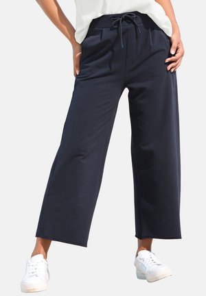 Woman wearing black wide-leg pants with drawstring waist and white sneakers, standing with hands on hips.