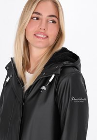 Čierna nepremokavá bunda s kapucňou lemovanou umelým kožušinou, s logom strieborného dáždnika a bielym šnúrkovým uzáverom. Hladká textúra so striebornými kovovými akcentmi.