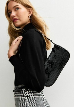 Borsa a tracolla in pelle nera con texture a stampa coccodrillo, forma rettangolare e tracolla sottile, abbinata a un maglione nero e pantaloni a scacchi.