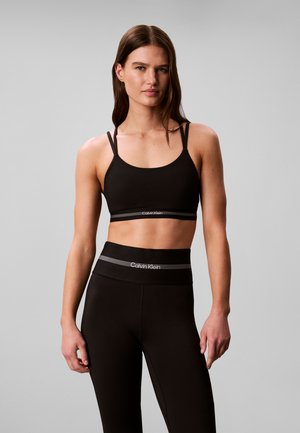 Vrouw met lang bruin haar die een zwarte Calvin Klein sport-bh en hoog getailleerde leggings draagt, staand tegen een effen grijze achtergrond.