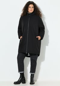 Sort knælængde frakke med lynl�ås foran og hætte, med sidelommer. Kombineret med mørke mønstrede jeans og sorte ankelstøvler.