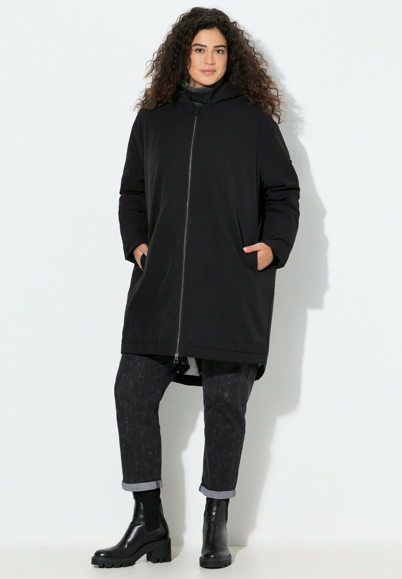 Sort knælængde frakke med lynlås foran og hætte, med sidelommer. Kombineret med mørke mønstrede jeans og sorte ankelstøvler.