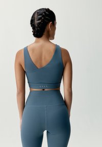 Teal športový crop top s dizajnom V-zadného dielu, ktorý má priliehavý strih a hladký materiál. Skombinovaný s legínami s vysokým pásom a textúrovaním.