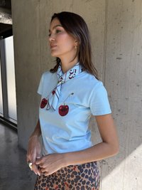 Světle modré tričko s krátkým rukávem se červenými grafikami třešní, doplněné bílým a burgundským šátkem s vzorem uvázaným na krku a tečkovanou sukní.